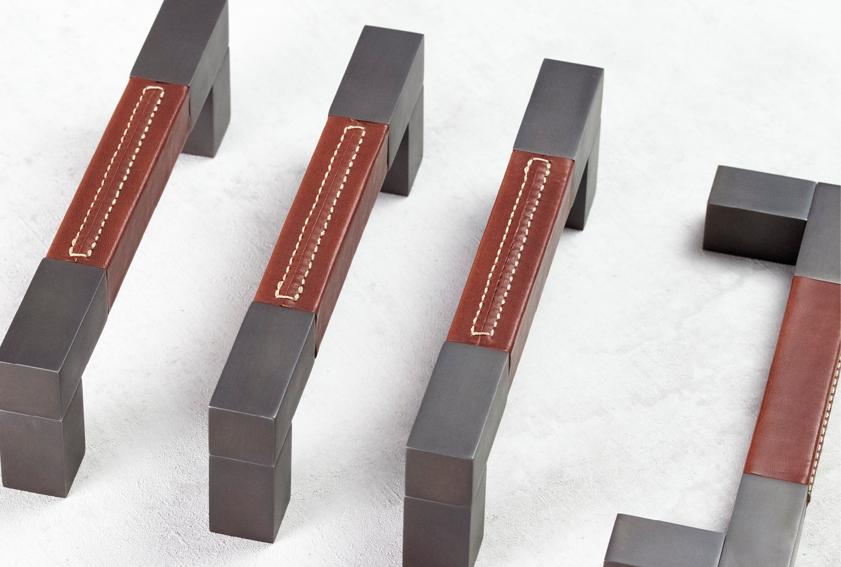 Image of four custom length recess leather square D pull handles. One of the pull handle is positioned on the side. Grip of the pull handle is finished in chestnut leather. The end caps and legs are finished in matt black chrome.