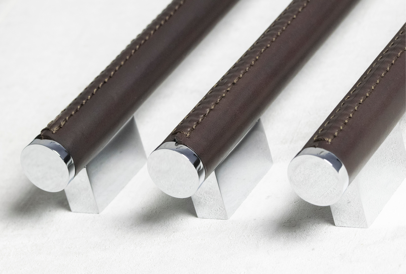 The image display, three coffin cabinet pull, positioned parrel to one another. The cabinet pull grip, is covered in a chocolate leather, joined with a hand stitch. Two small rectangular legs are positioned either end of the cabinet pull.