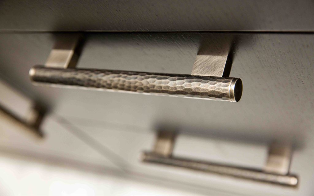 Hand hammered coffin leg scroll cabinet handle on a dark wooden drawer unit. Finished in Burnished brass, a dark brown living finish which will patinate over time and use.