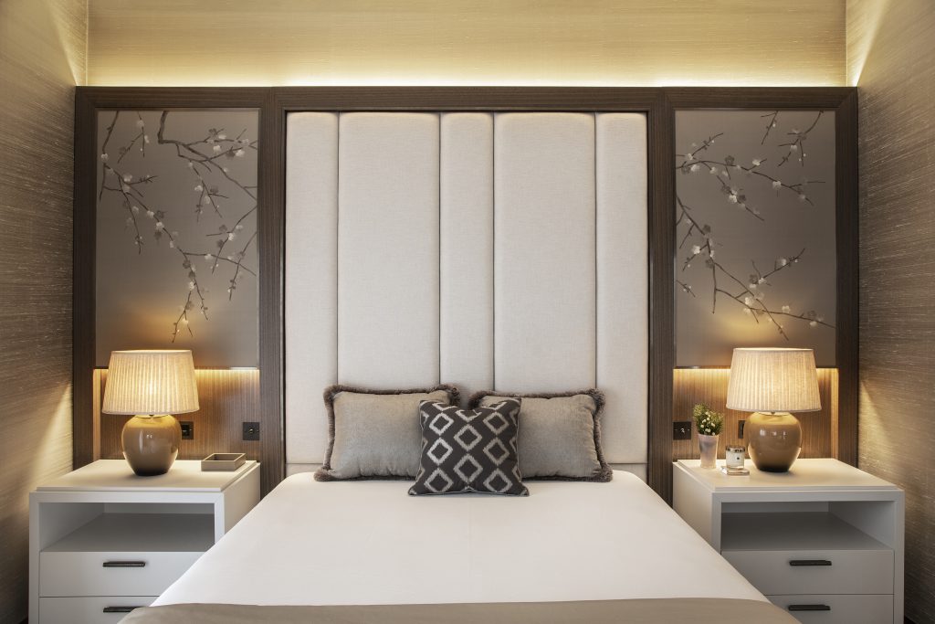 Hand hammered brass cabinet handles in burnished nickel, a moody grey textured finish. They sit on two white, two drawer bedside tables either side of the large king bed. Neutral tones and muted colours have been used in this bedroom decor with floral motif wallpaper to accentuate the colour pallet. Matching lamps with mustard yellow bases on top of both bedside tables. Scatter cushions are propped up against a large built in head board which accent the colours of the surrounding wallpaper and room.