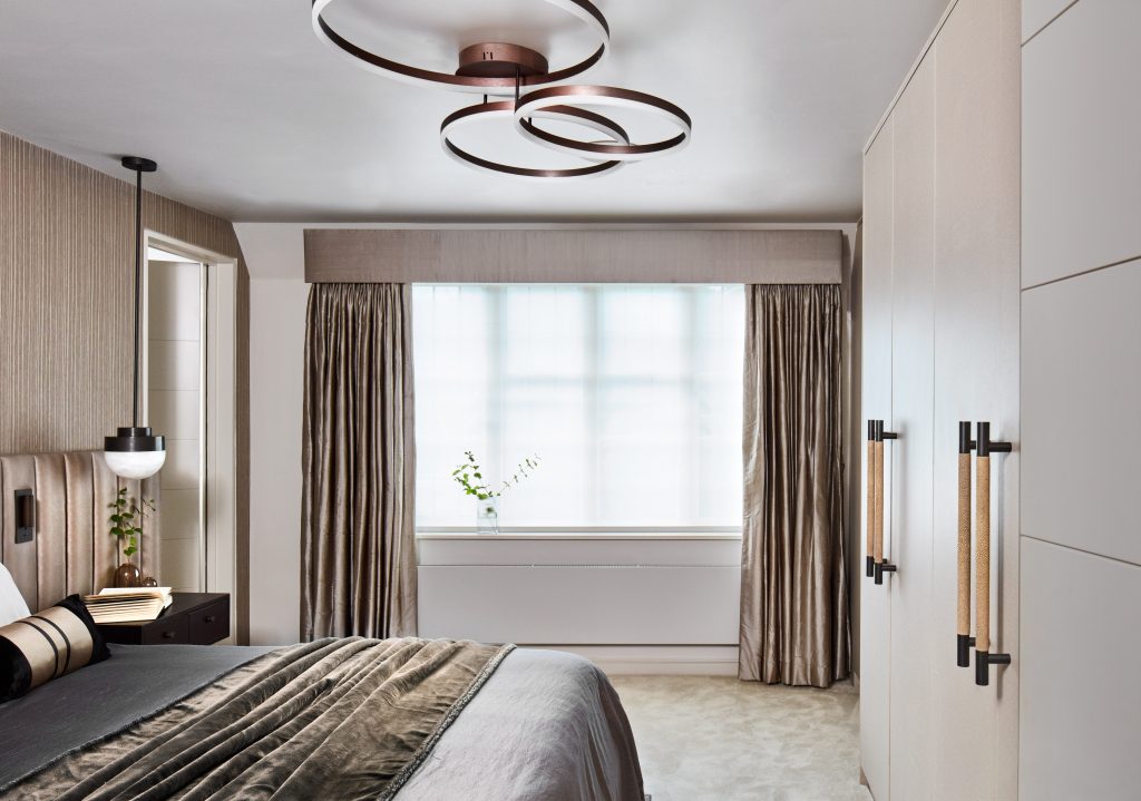 A muted decor styled bedroom with deep browns and dark grey purples. four strong and bold large shagreen door pulls on the wardrobes at the foot of the bed stand out. The shagreen texture, like that of a sting ray skin, is a sandy colour makes up the grip of these pull handles and sits against a matt black chrome metal legs. Circular statement ceiling lights sit centrally over the bed.