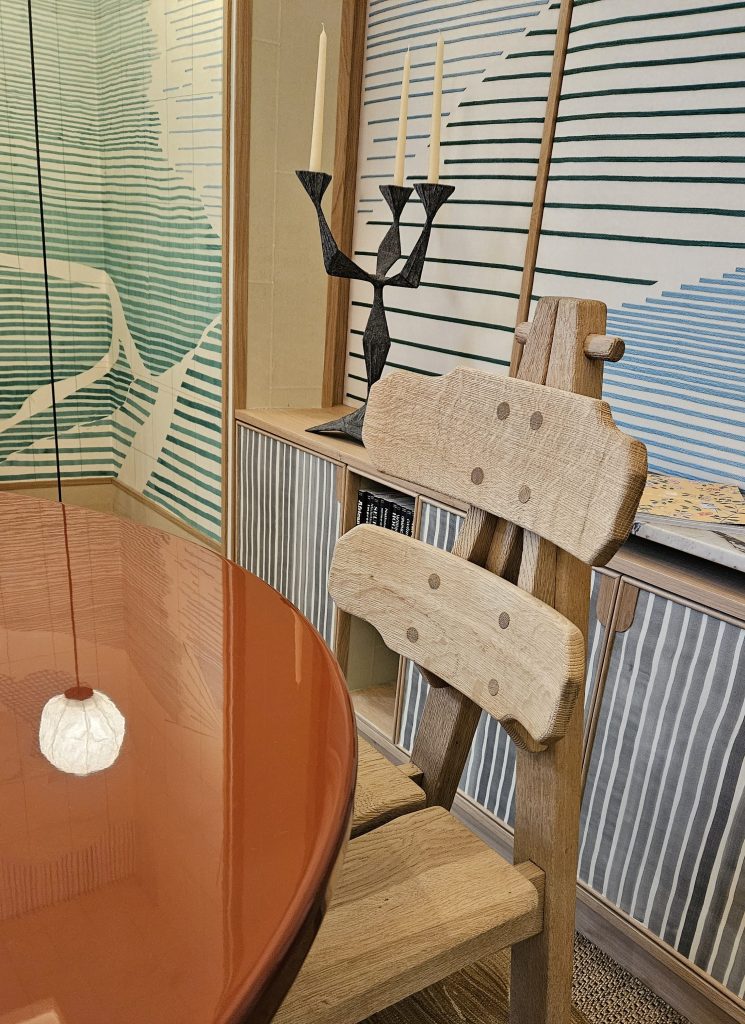 De Gournays Morning Room, an abstract space designed arund weather, nature and architecture. Hand painted wall coverings and light airy colours are used. A burnt orange table with bespoke wood chair is the central part of the image with the wallpaper and lighting in the background.