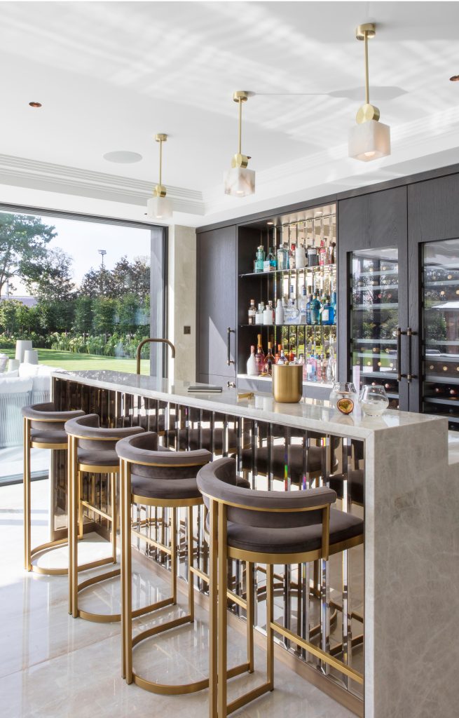 A luxury drinks bar situated in a vast new extension onto a large Tudor house. Decorated glass and marble surface creates the central bar area with art deco style bar stools. In the background a large collection of spirits sit on the floating shelves.