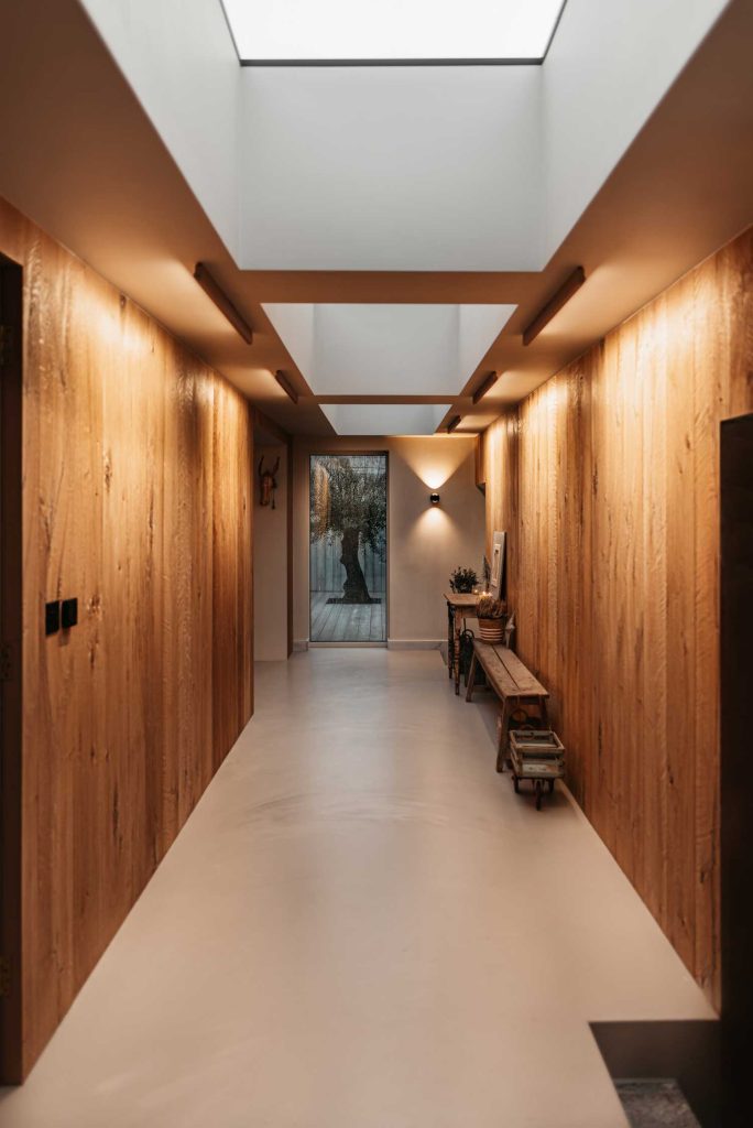 Long wooden panelled entrance hall of a modern refurbished kitchen. Light granite flooring and warm orange lighting creates a warm, inviting feel.