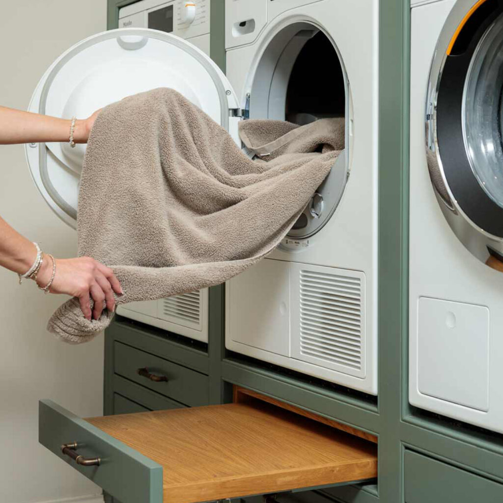 A person removing a towel from a integrated tumble dryer in this luxury utility room. Bespoke bottom slide out drawers built to rest laundry or baskets on, fixed with Turnstyle Designs leather cabinet handle.