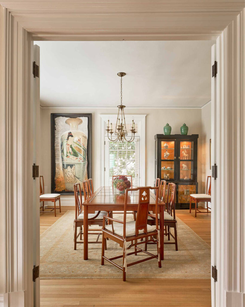 A large dining room of a North Carolina house. Large paintings on the walls and a central dining table sits in th middle on a large decorative rug and wooden floor. Looking through a double door entrance way into this dinning room.