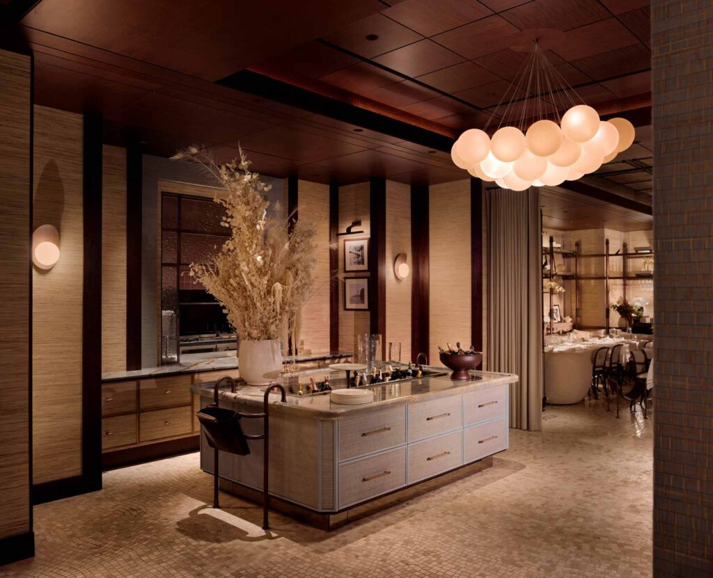 Sophisticated restaurant interior featuring a central island with drawers and brass hardware, styled with glassware and a large arrangement of dried botanicals in a ceramic vase. The walls are clad in textured neutral-toned panels with dark wood accents and framed black-and-white photographs. A sculptural chandelier made of clustered, glowing orbs hangs overhead, casting a warm, ambient light. In the background, a refined dining area with elegant seating and curated shelving is visible through an open curtain, maintaining a seamless flow of the luxurious, earthy-toned design.