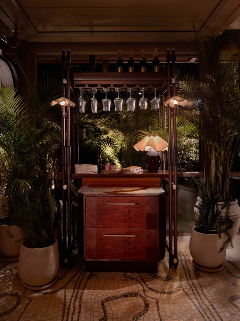 A mini bar section from a fine dining restaurant, hidden by atmospheric lighting and plants, the dark wooden mini bar uses decorative hardware with a Shagreen texture for its cabinet handle. This handle is handmade by Turnstyle Designs.