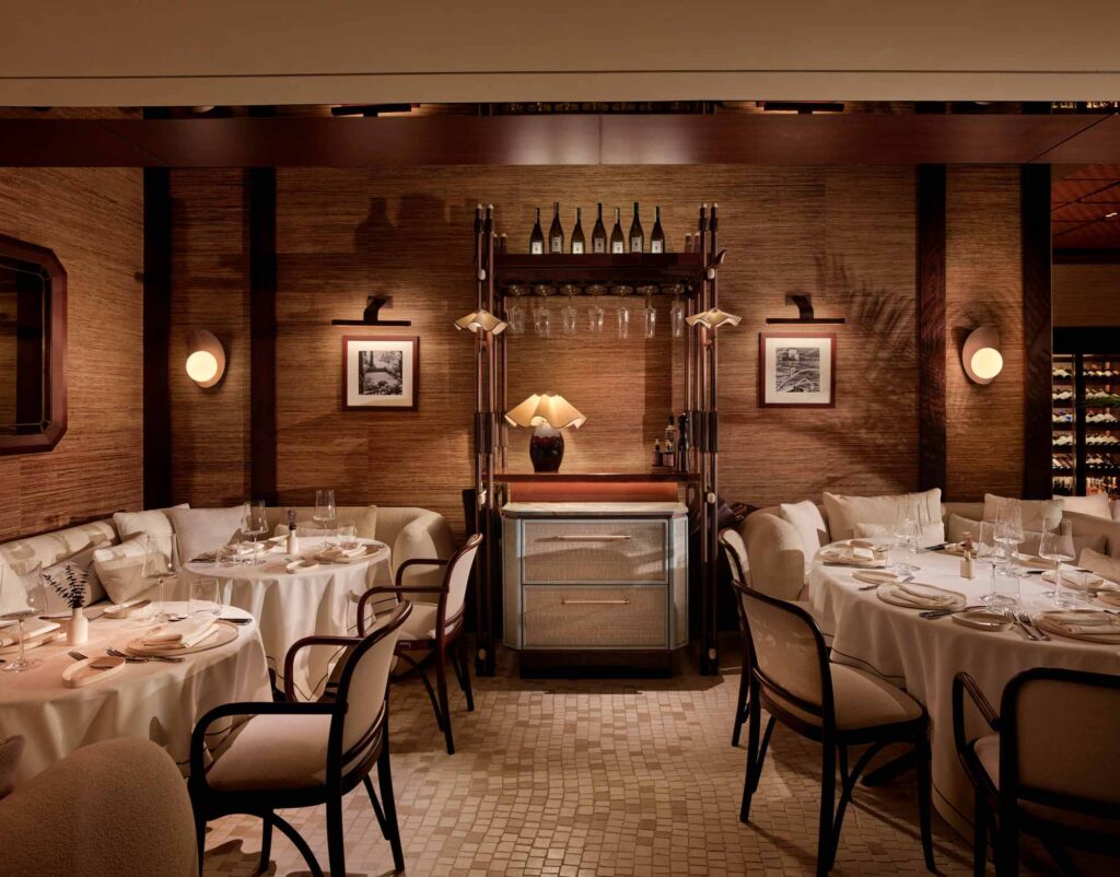 Elegant, softly lit restaurant interior with a round table set for fine dining. The space features neutral tones, natural wood chairs with canvas backs, plush upholstered seating, and curved glass walls with sheer curtains. A mosaic floor and potted greenery add texture and warmth, while shelves of glassware and wine bottles enhance the luxurious ambiance. The overall design evokes refined comfort and understated sophistication.