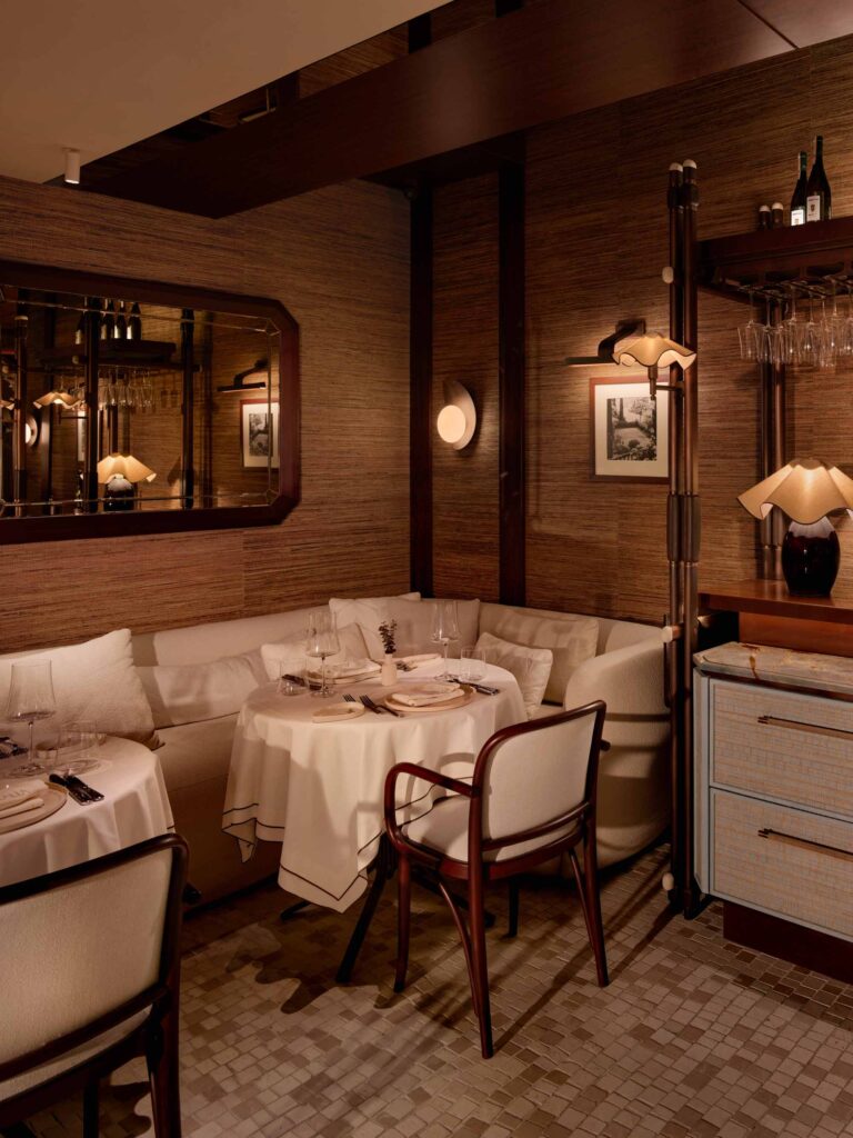 Sophisticated restaurant interior clad in textured neutral-toned panels with dark wood accents and framed black-and-white photographs. In the background, a refined dining area with elegant seating and curated shelving is visible.