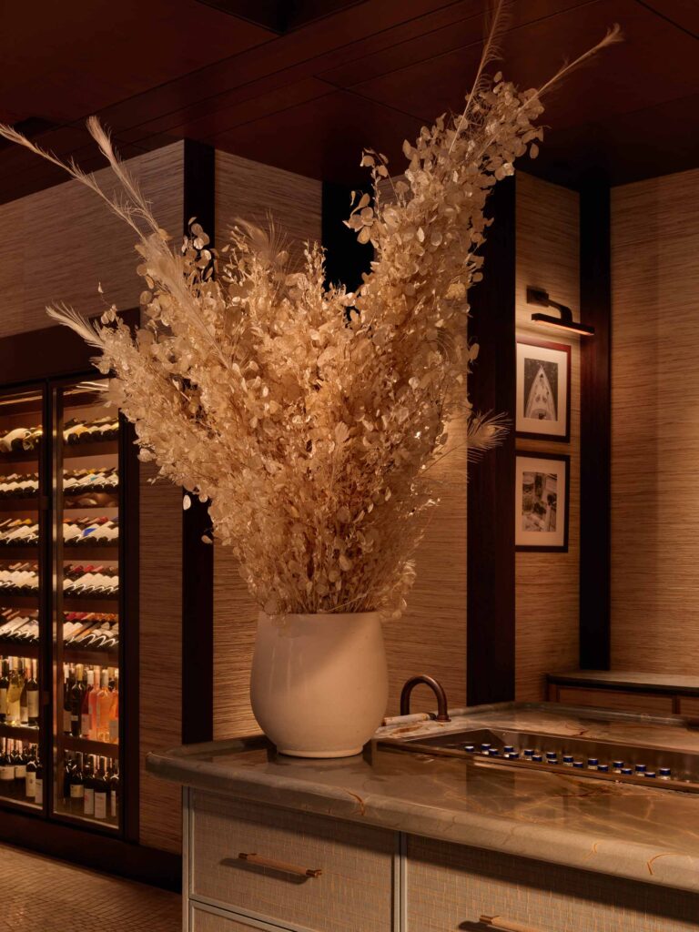 Close-up of a luxurious restaurant setting featuring a large, textured ceramic vase filled with dried golden foliage and feathers, placed on a richly veined marble countertop. The island beneath showcases finely detailed drawers with shagreen-patterned fronts and brass pulls. In the background, a softly lit wine fridge displays an extensive collection of bottles. Warm ambient lighting and natural materials like wood and grasscloth wall coverings create a refined, organic elegance.