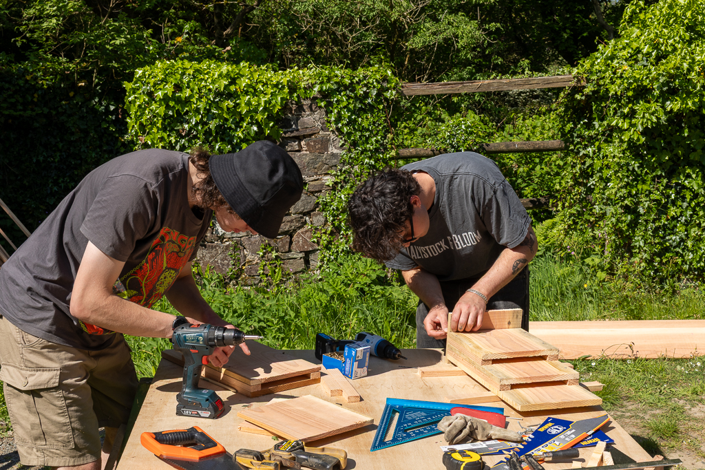 Two people using tools are creating bat boxes for a local organisation. They are holding drills and saws.