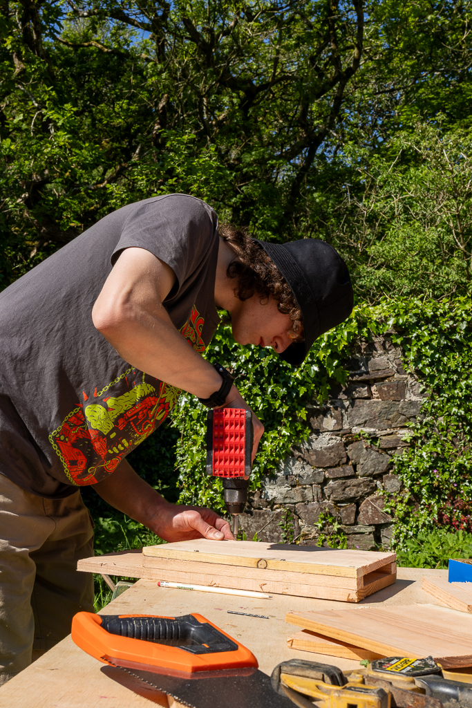 A person is holding a drill on a work bench and screwing holes into cut bits of wood to make bat boxes. These bat boxes are for the National trust to erect around the peppercombe area.