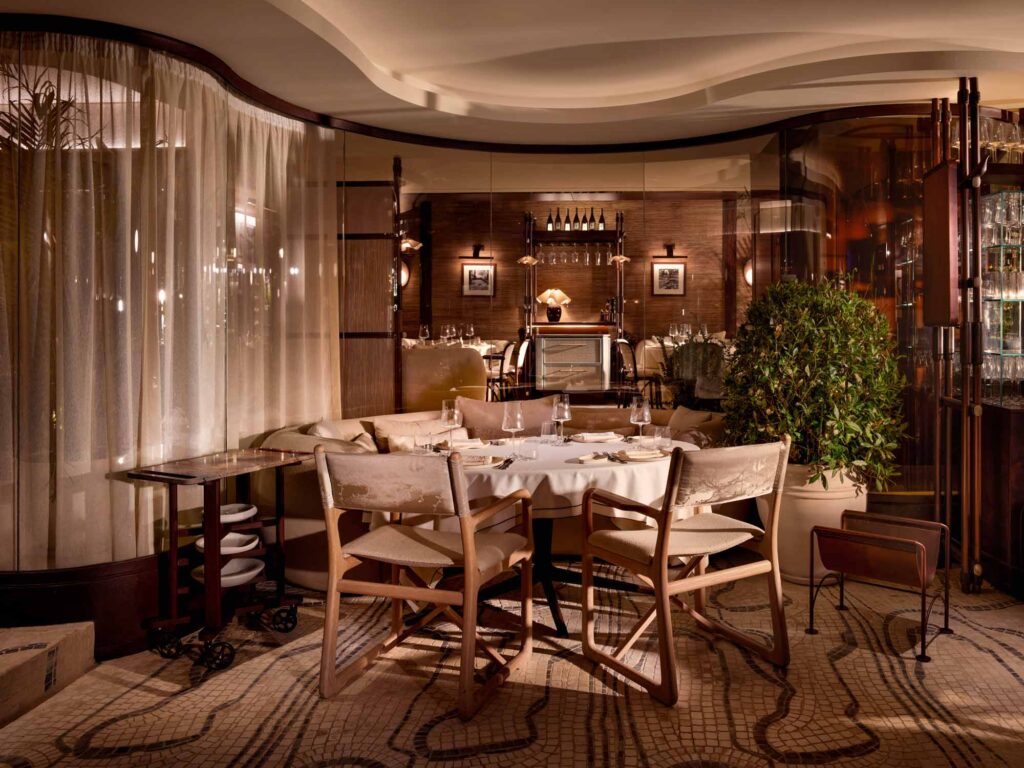 Elegant, softly lit restaurant interior with a round table set for fine dining. The space features neutral tones, natural wood chairs with canvas backs, plush upholstered seating, and curved glass walls with sheer curtains. A mosaic floor and potted greenery add texture and warmth, while shelves of glassware and wine bottles enhance the luxurious ambiance. The overall design evokes refined comfort and understated sophistication.