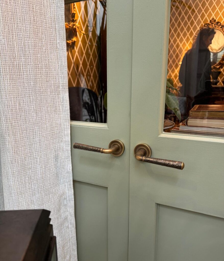 Image shows double doors with matching Tube Goose Neck door levers by Turnstyle Designs. These levers are hand hammered and finished in a Burnished Brass finish. Featured on the courtyard room at the 2025 WOWhouse exhibition in London's chelsea harbour.