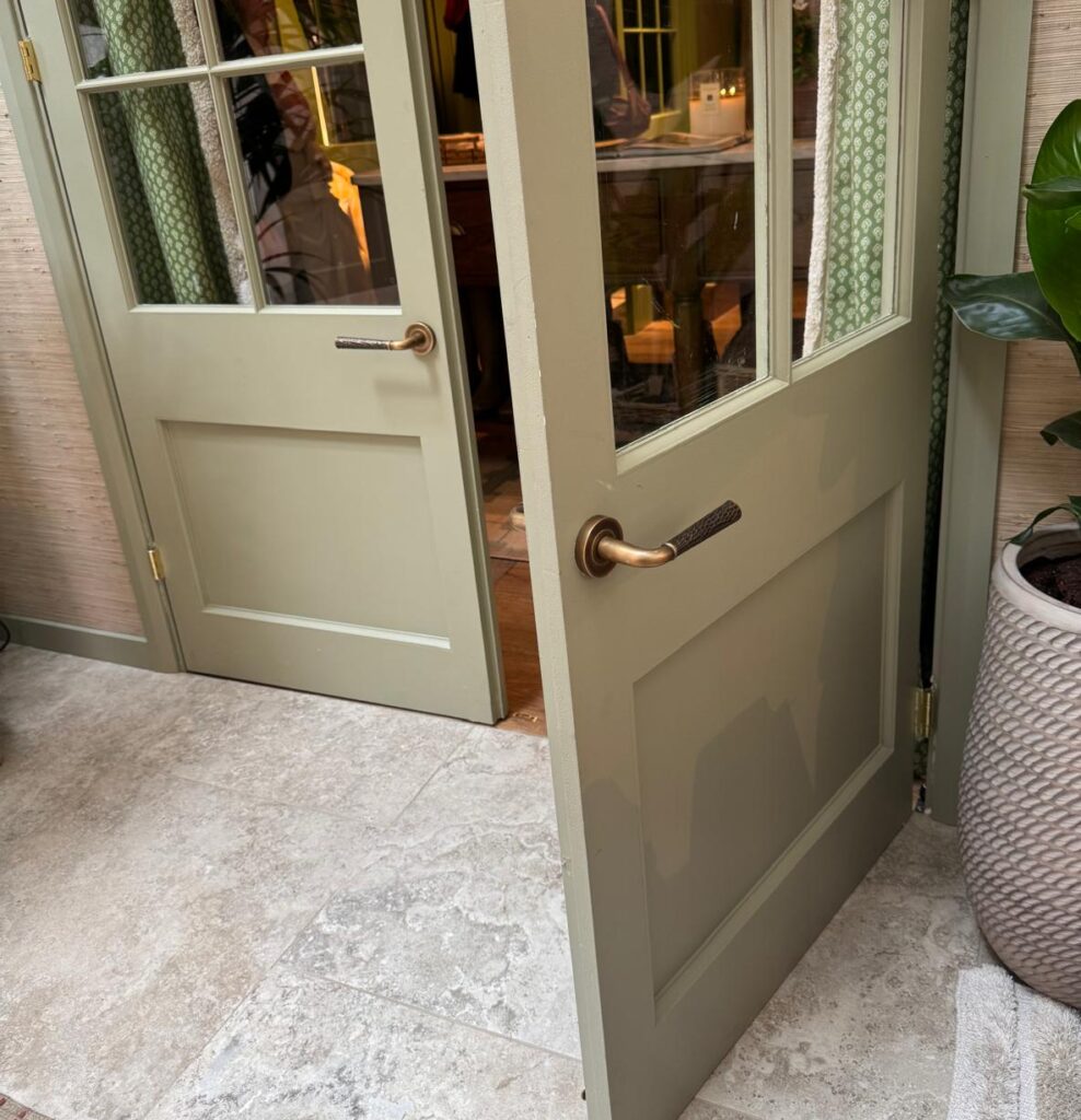 Image shows a set of double doors with one open, highlighting the hand hammered brass door lever on them. These door levers are made by Turnstyle Designs and have been used on this exhibition space at WOWhouse in the Design Centre at Chelsea Harbour.