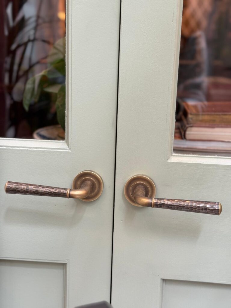 Two hand hammered door levers shown on pale sage green double doors that are closed. The doors have glass windows allowing the viewer to see into the room that is a dark warm amber colour with moody lighting.