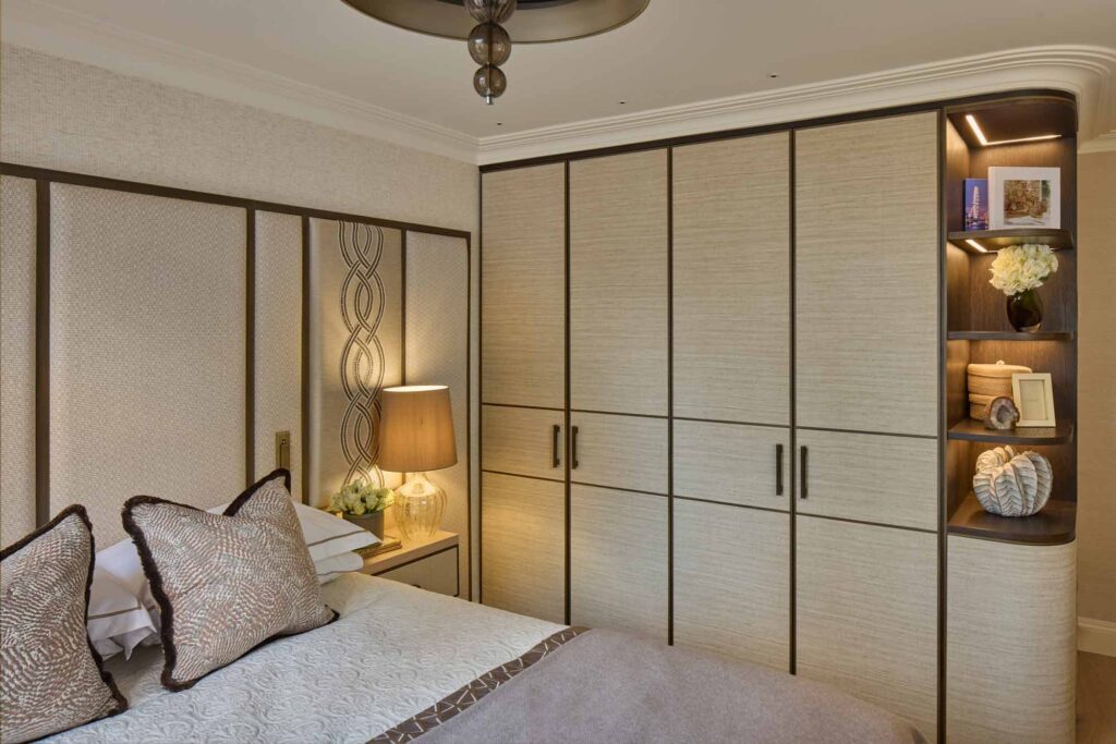 A luxurious bedroom featuring a bespoke fitted wardrobe with light wood panels framed in dark trim, integrated open shelving with accent lighting, and decorative accessories including books, flowers, and sculptural objects. A neatly made bed with textured pillows and a quilted throw sits beside a nightstand with a glass lamp, all in warm neutral tones