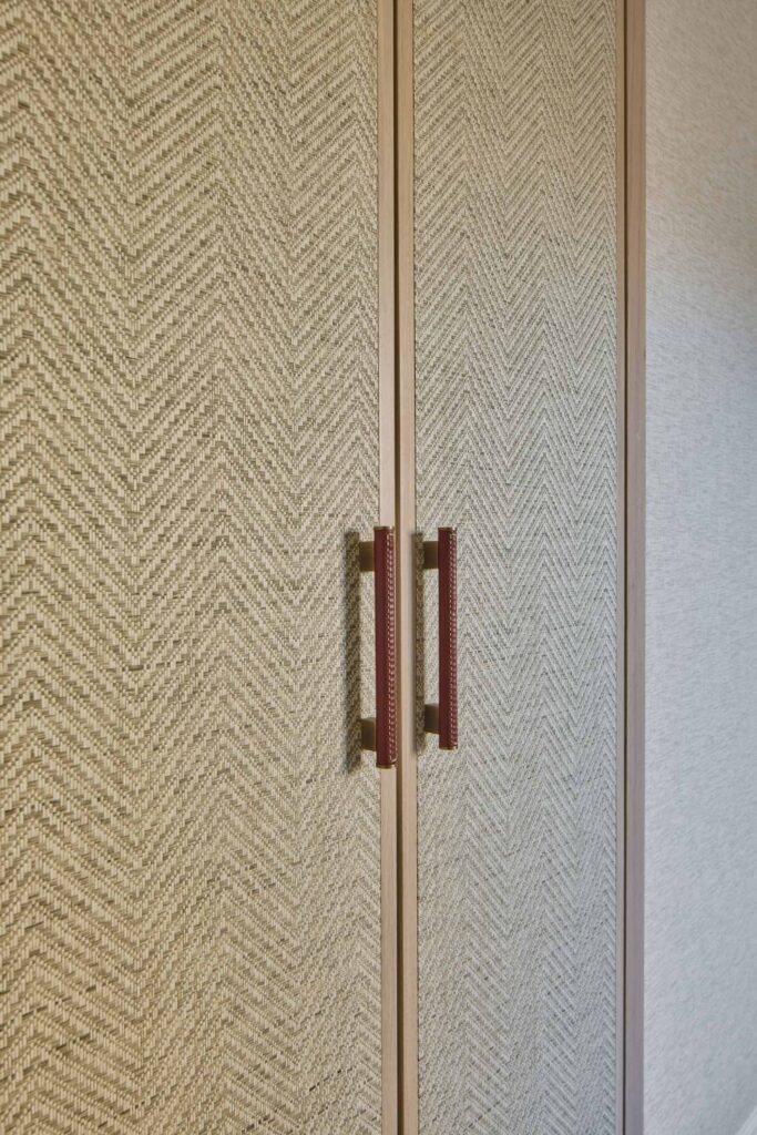 Close-up of a wardrobe with herringbone-patterned textured panels in soft neutral tones, framed in light wood, featuring elongated handles wrapped in rich Chestnut leather with Stitched detail by Turnstyle Designs, alongside woven textures and gold-tone accents