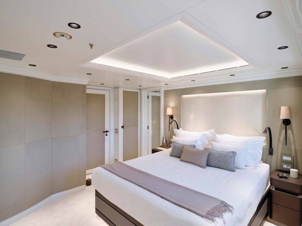 "Luxurious, modern bedroom with a neutral palette featuring a king-sized bed dressed in crisp white linens, layered with decorative pillows and a soft throw. The walls are upholstered in soft beige panels, complemented by recessed lighting in the ceiling and subtle wall sconces. Minimalist nightstands flank the bed, each with contemporary black reading lights. The space is bright, serene, and meticulously detailed."