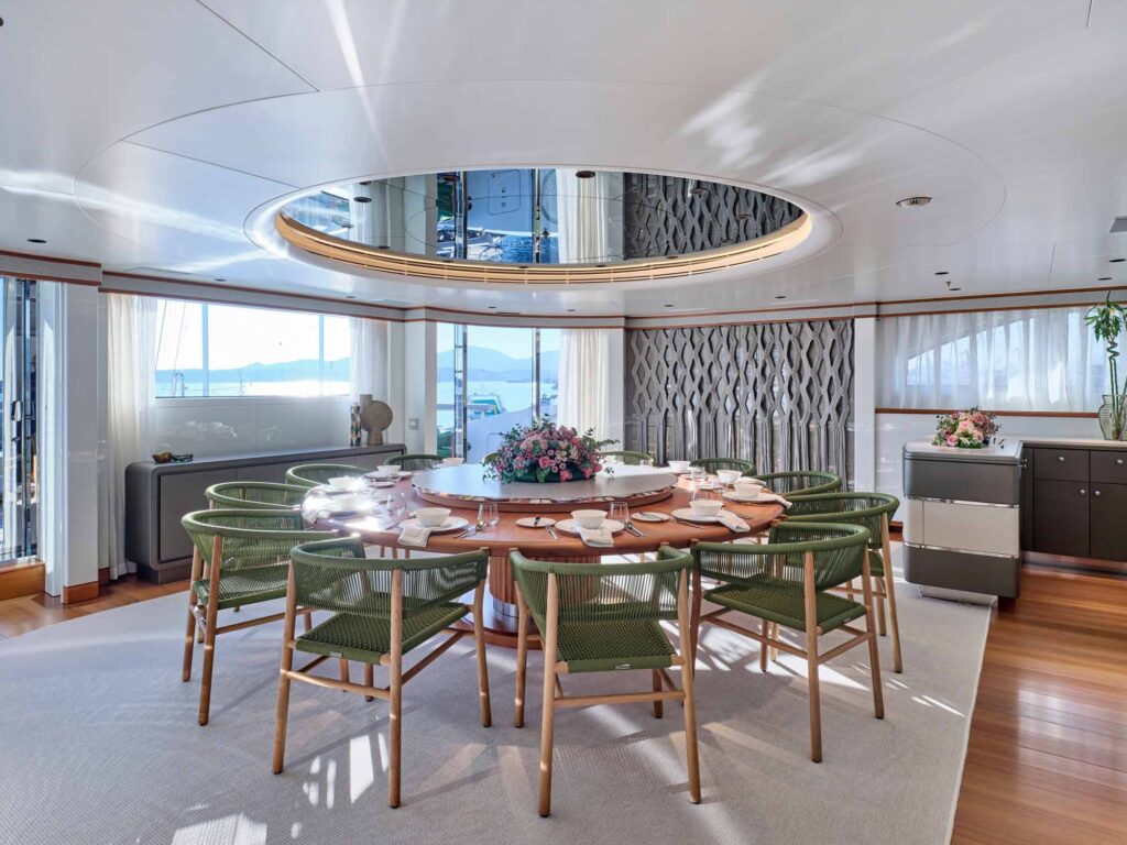 Elegant superyacht sky lounge dining area with a circular wooden table set for eight, green woven chairs, fresh floral centrepiece, and panoramic sea views through large windows, accented by natural light and a mirrored ceiling feature.