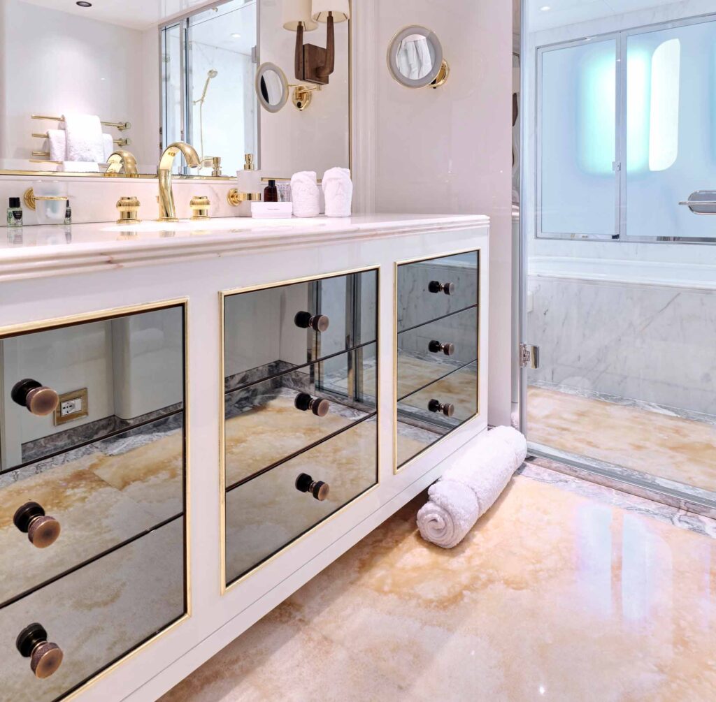 Luxurious yacht bathroom with polished marble flooring in warm amber tones, mirrored vanity drawers framed in gold, and silver-bronze cabinet knobs. The space features a large backlit mirror, gleaming gold fixtures, and a walk-in shower with frosted glass doors, creating an elegant and sophisticated atmosphere.
