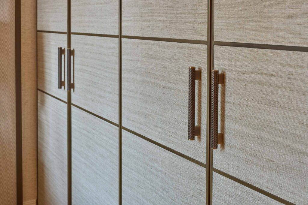 Close-up of a fitted wardrobe with textured light wood panels framed in dark trim, featuring elongated vertical handles in a woven amalfine finish by Turnstyle Designs in Silver Bronze with Fine Antique.