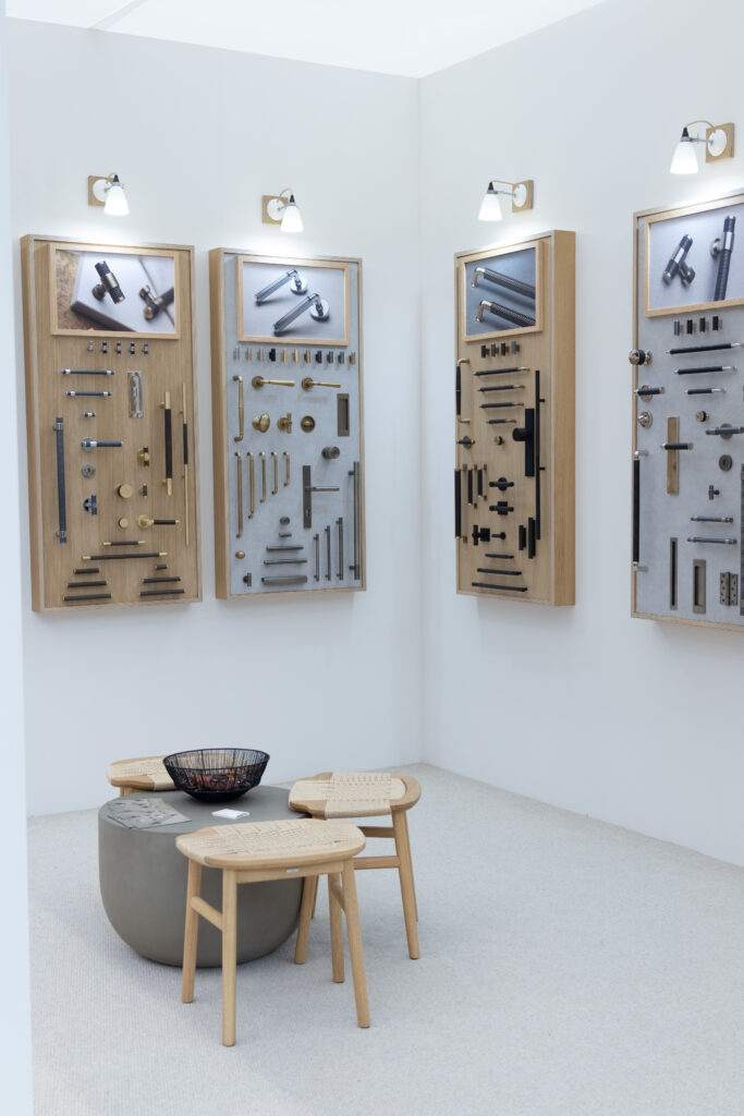 A shot of a corner of Turnstyle Designs' event stand: oak display boards featuring luxury door and cabinet hardware are mounted on white walls. In the foreground - a seating area with a small table and three short stools.