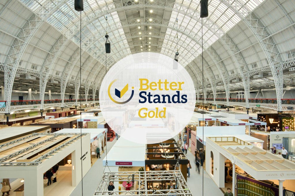 A photo overlooking many exhibition stands under a large, barrel vaulted roof with glass canopy. The centre of the image is overlaid with a logo which reads Better Stands Gold