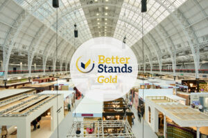A photo overlooking many exhibition stands under a large, barrel vaulted roof with glass canopy. The centre of the image is overlaid with a logo which reads Better Stands Gold